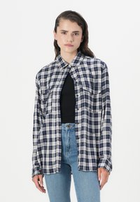 Mujer joven con cabello largo castaño, vistiendo una camisa de cuadros azul marino y blanca desabrochada sobre una blusa negra y unos jeans de cintura alta azules.