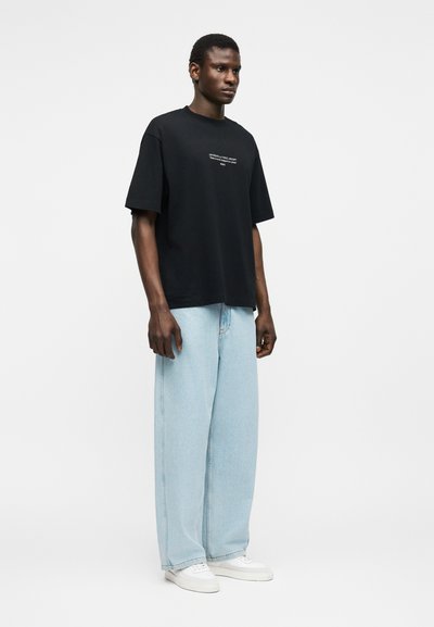 Άνδρας που φοράει μαύρο φαρδύ T-shirt με μικρό λευκό κείμενο, ανοιχτό μπλε τζιν με φαρδιά πόδια και λευκά αθλητικά παπούτσια, στέκεται μπροστά σε απλό φόντο.