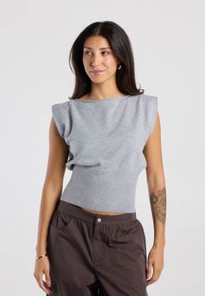 Frau mit langen dunklen Haaren, die ein ärmelloses graues Stricktop und braune Hose trägt und ein florales Tattoo am linken Unterarm zeigt.