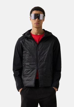 Mand iført store reflekterende solbriller, sort quiltet hættejakke, rød skjorte under og sorte bukser, stående med hænderne i lommerne.