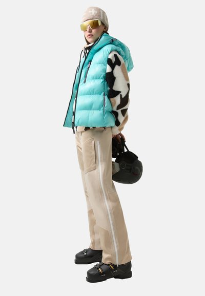 Een donzen bodywarmer in turquoise, een gebreide trui met patroon, beige skibroek met zijritsen en zwarte skischoenen, met een zwarte helm in de hand.