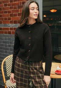 Woman wearing a buttoned dark brown cardigan and brown plaid pants, standing near a café table with a cup of coffee and pastry.