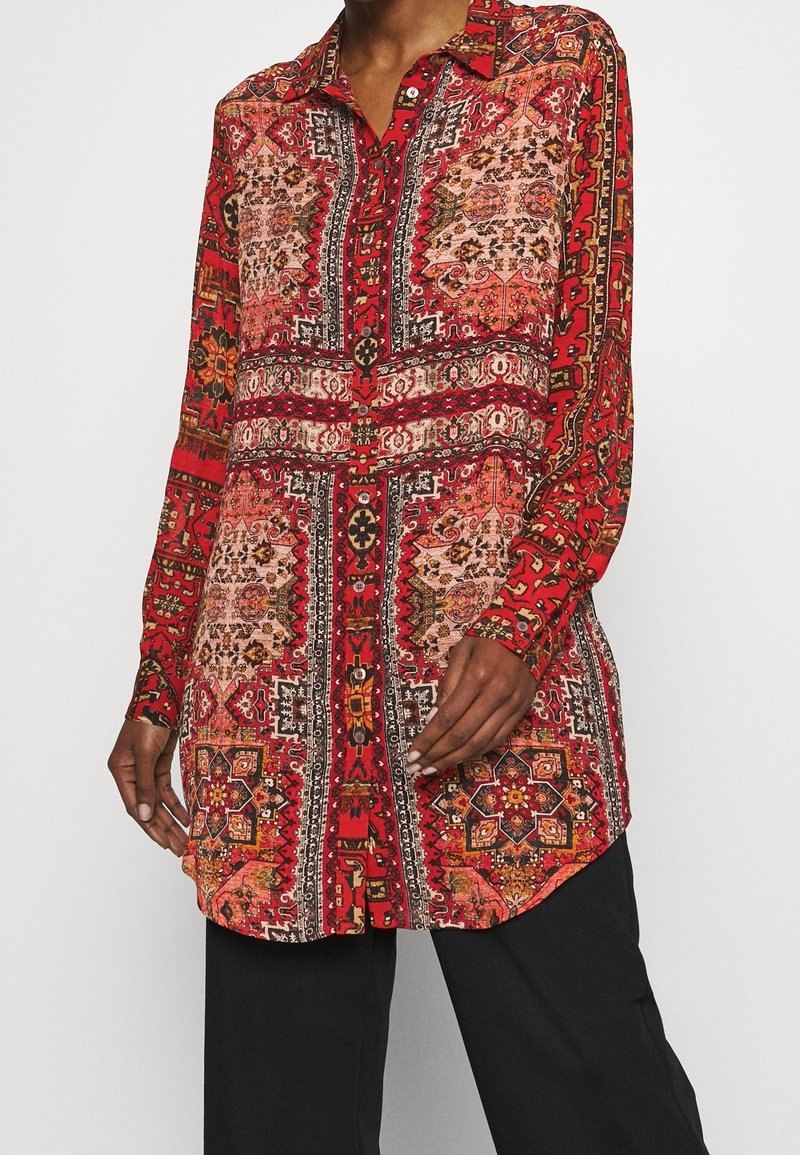 Personne portant une tunique rouge à manches longues avec des motifs géométriques et floraux, associée à un pantalon noir, debout devant un fond uni.