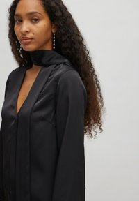 Schwarze Satinbluse mit tiefem V-Ausschnitt, hohem Kragen und langen Ärmeln. Verfügt über eine glatte Textur und dezente Vordernahtdetails.