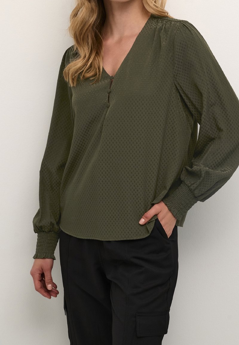 Blouse vert olive avec col en V, motif texturé, manches longues bouffantes et poignets élastiques. Présente un détail de bouton au niveau de l'encolure.
