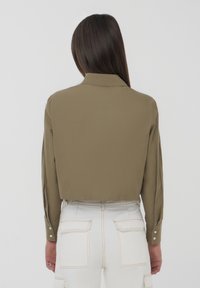Camicia verde oliva tagliata con colletto, maniche lunghe e polsini con bottoni. La camicia presenta una superficie liscia ed è abbinata a pantaloni bianchi.