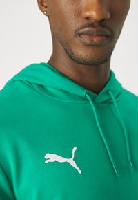 Sweatshirt verde feito de tecido macio, com bolso canguru. Apresenta o logótipo branco da Puma no peito e cordões na capuz.