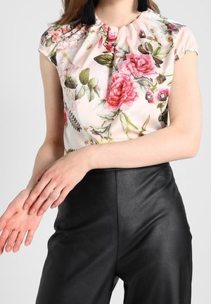 Frau trägt eine kurzärmelige Bluse mit Blumenmuster und schwarze hoch taillierte Lederhose, die Hände berühren sanft ihren linken Unterarm.