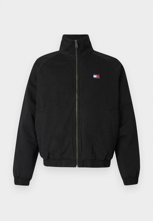 Veste bomber noire avec col montant, fermeture éclair avant, poignets et ourlet élastiques, présentant un petit logo rouge, blanc et bleu sur la poitrine.
