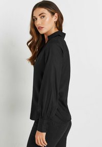 Chemise noire à boutons avec col, manches longues bouffantes et texture lisse, présentée de profil sur un fond neutre.