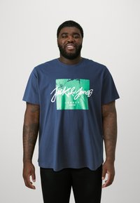 Navy blue cotton t-shirt with short sleeves. Features a rectangular green and white graphic with "Jack & Jones" text and "Legacy" below.