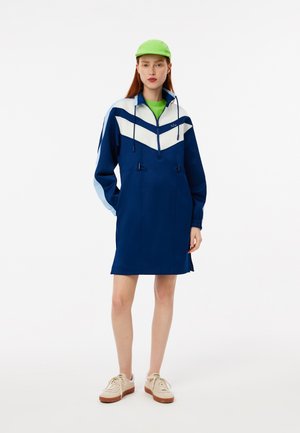 Marineblauwe hoodie jurk met witte en lichtblauwe chevronaccenten, voorzien van trekkoorden en een voorkeurszak. Gecombineerd met een groene muts en sneakers.