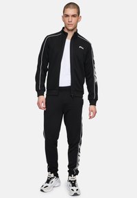 Chándal deportivo negro con chaqueta de cremallera, rayas de acento blancas y detalles de logo. Los pantalones tienen un corte ajustado y puños elásticos.
