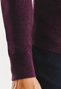 Męska ręka w ciemnofioletowym swetrze dzianym z prążkowanymi mankietami, pokazana na jasnym tle w ciemnych spodniach.