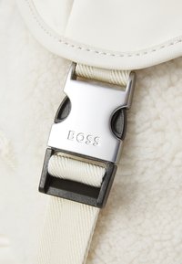 A silver metal buckle with the text "BOSS" on a cream-colored strap, attached to a fluffy textured fabric in a light beige shade.