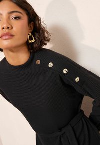 Žena s vlasy po ramena a tmavou barvou nosí zlaté geometrické náušnice a černé šaty s vroubkováním, které mají na rameni čtyři zlaté knoflíky.