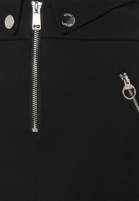 Veste en tissu noir avec une fermeture éclair à l'avant et une poche zippée sur le côté. Des accents en métal argenté rehaussent le design épuré et la texture.