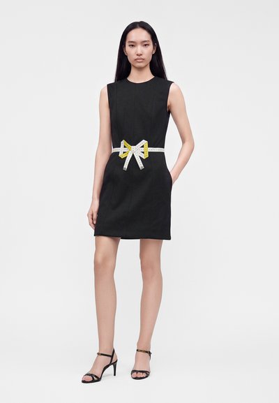 Femme en robe noire sans manches avec un mètre ruban blanc et jaune noué en nœud à la taille, portant des sandales noires à talons hauts.