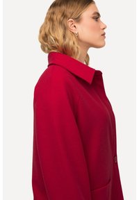 Manteau en laine rouge avec un large col, des poches latérales et une fermeture à boutons. Le tissu semble lisse avec une finition texturée.