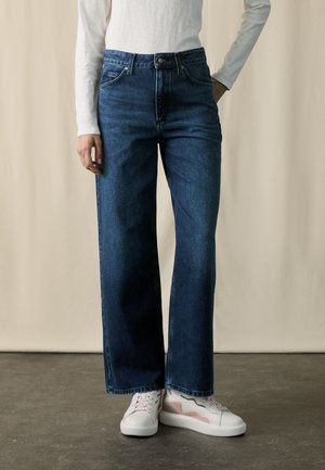 Vrouw die een blauw hoogwaterbroek met brede pijpen draagt, een witte top met lange mouwen en witte sneakers met roze en grijze accenten, staand op een grijze vloer.