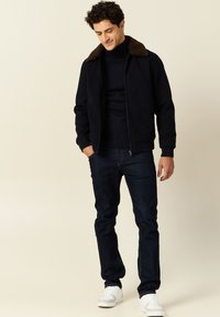 Czarna kurtka bombowa z brązowym futrzanym kołnierzem, ciemny golf, ciemne dżinsy i białe trampki. Model stoi w zrelaksowanej postawie.