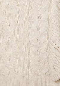 Tissu tricoté de couleur crème avec des motifs verticaux en câble et en côtes, montrant des points texturés et les détails d'une matière en laine douce.