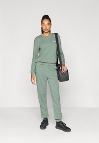 Zielona bawełniana bluza i pasujące spodnie dresowe z okrągłym dekoltem i mankietami, zestawione z czarną torbą na ramię i czarnymi sportowymi butami.