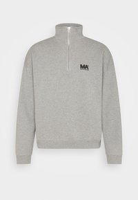 Sudadera gris de cuello alto hecha de tela suave, con cremallera frontal hasta la mitad y un logo negro "MA" en el pecho izquierdo. Puños y dobladillo acanalados.