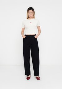 Witte geribbelde crop top met rode hartdetails, gecombineerd met hoge zwarte wijde broek en kastanjebruine instappers. Zachte texturen.