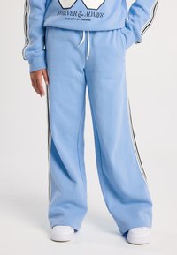 Henkilö, jolla on päällä vaaleansiniset leveälahkeiset verkkarit, joissa on mustavalkoiset sivusuunnassa kulkevat raidat, sekä valkoiset lenkkarit. Hän seisoo käsi taskussa.