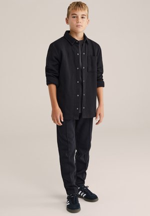 Cămașă neagră cu nasturi și buzunar, purtată peste un tricou negru. Asortată cu pantaloni sport negri și adidași negri cu dungi albe.