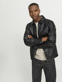 Chaqueta de cuero negra con diseño de cuello, cierre de cremallera y bolsillos frontales, combinada con un suéter gris claro y jeans oscuros.