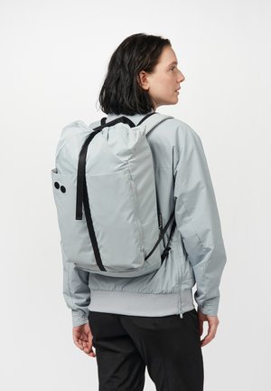 Hellgrauer Rucksack mit Zugbandverschluss, schwarzen Trägern und zwei runden Akzenten. Hergestellt aus einem glatten, leichten Material.