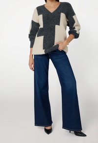 Pull surdimensionné à col en V gris et crème avec un motif géométrique, associé à un jean large bleu foncé et des talons noirs.