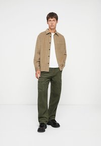 Camisa de cuadros marrón con botones, camiseta blanca, pantalones cargo verde oliva y zapatos negros gruesos. Corte recto con bolsillos en los pantalones.