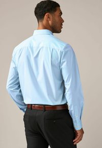 Camisa de manga larga azul claro con un cuello rígido, tela suave y detalles de botones en los puños, combinada con pantalones oscuros y un cinturón marrón.