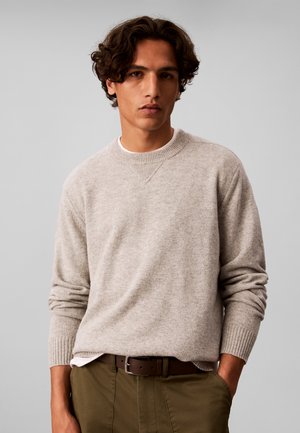 Jonge man met krullend haar die een lichtgrijze trui met ronde hals en olijfgroene broek met een bruine riem draagt, staand tegen een effen grijze achtergrond.