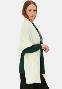 Kołdra w kolorze kości słoniowej, wykonana z miękkiego materiału, prostokątna, z frędzlami na brzegach, noszona na ciemnozielonym swetrze. Gładka tekstura, lekka konstrukcja.