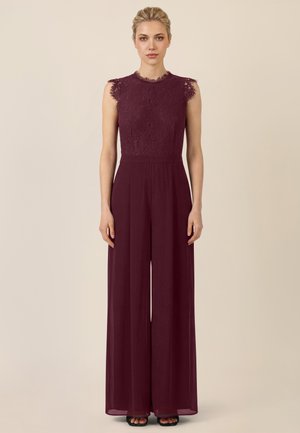 Frau in ärmellosem, burgunderfarbenem Spitzen-Jumpsuit mit weitem Hosenbein, stehend vor einem schlichten beigen Hintergrund.
