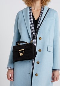 Musta mokkanahkainen käsilaukku kultaisilla yksityiskohdilla, suorakulmainen muoto, yläkahva ja irrotettava hihna, joka on käytössä vaalean sinisen takin kanssa.