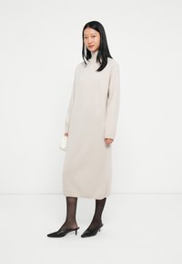 Vrouw die staat in een beige midi-jurk met lange mouwen en col, zwarte doorschijnende panty’s, zwarte kittenheels, en een kleine witte clutch.