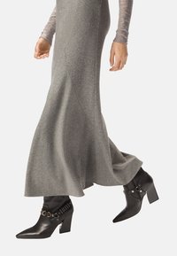 Vestido largo y fluido de color gris con la parte superior ajustada. Botines negros hasta el tobillo con punta puntiaguda, tacón grueso y detalles de anillos dorados.