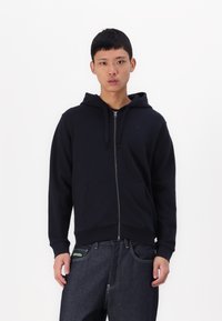 Mornarska zip-up kapuca z vrvico za nastavljanje, sprednjimi žepi in rebrastimi manšetami. V kombinaciji s temnimi kavbojkami z zeleno oznako.