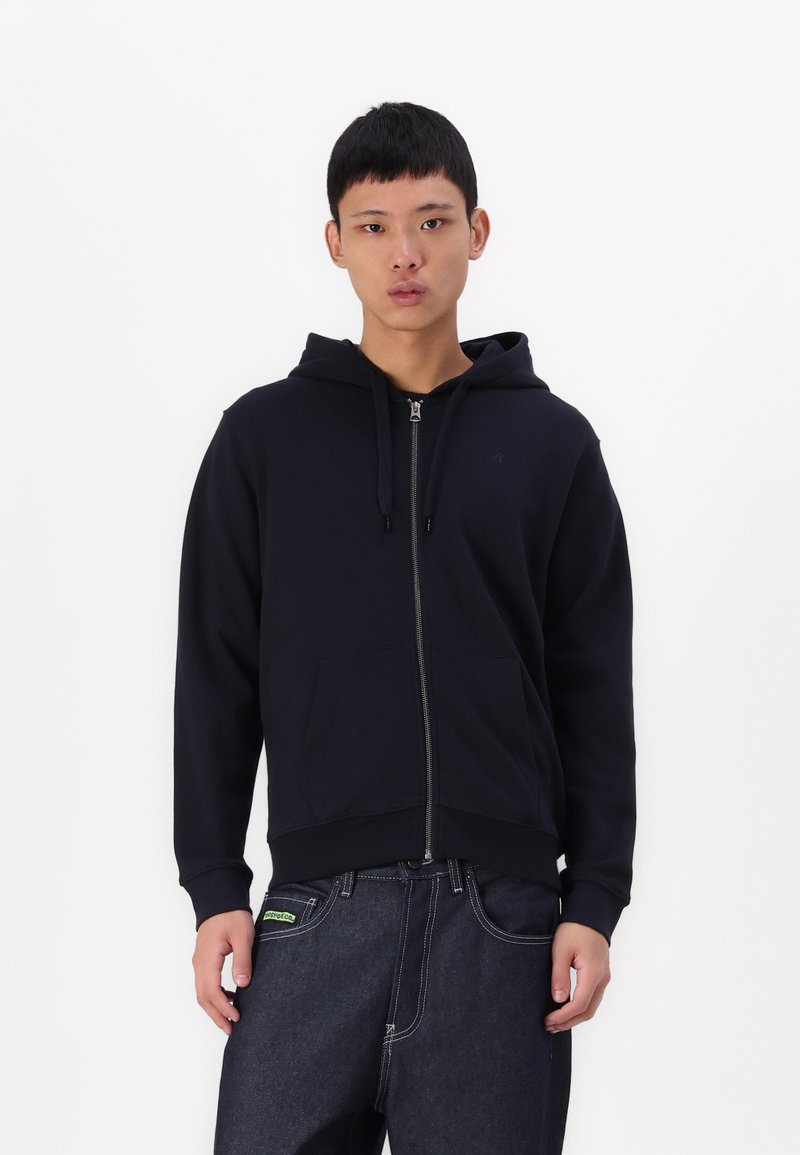 Mornarska zip-up kapuca z vrvico za nastavljanje, sprednjimi žepi in rebrastimi manšetami. V kombinaciji s temnimi kavbojkami z zeleno oznako.