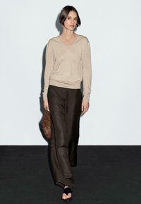 Das Modell schreitet selbstbewusst in einem beigen Pullover und dunklen Hosen, während es eine strukturierte braune Clutch hält, vor einem schlichten Hintergrund.