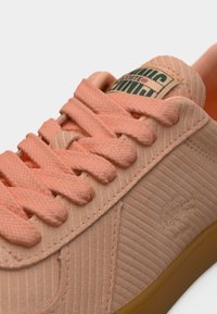 Ανοιχτό ροζ αθλητικό παπούτσι με υφασμάτινη υφή, λεπτά κορδόνια και λαστιχένια σόλα. Διαθέτει ετικέτα λογότυπου σε πράσινο και λευκό στο πλάι.