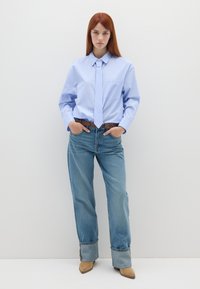 Himmelblau gestreifte Button-Down-Bluse mit einer passenden Krawatte, kombiniert mit hellblauen Jeans und beigen Absatzstiefeln. Das Modell steht mit den Händen in den Taschen.