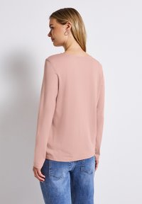 Långärmad, rundhalsad topp i mjuk rosa tyg, bärs med blå jeans. Slät textur, minimalistisk design och inga synliga mönster eller metalldelar.