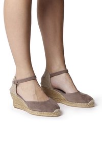 Hnědé semišové espadrilky na klínu s tkanou patou, kotníkovým popruhem a zaoblenou špičkou. Texturované tkaninové detaily na patě dodávají ozdobu.