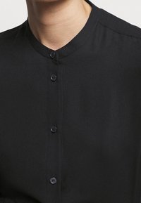 Chemise noire à col montant, avec un devant à boutons, une texture lisse et des boutons noirs espacés régulièrement.